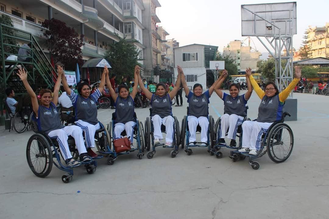 In the foreground of a basketball court are seven women in wheelchairs. They are stretching their arms up in the air and holding hands. The women are wearing jerseys and trainer shorts.
