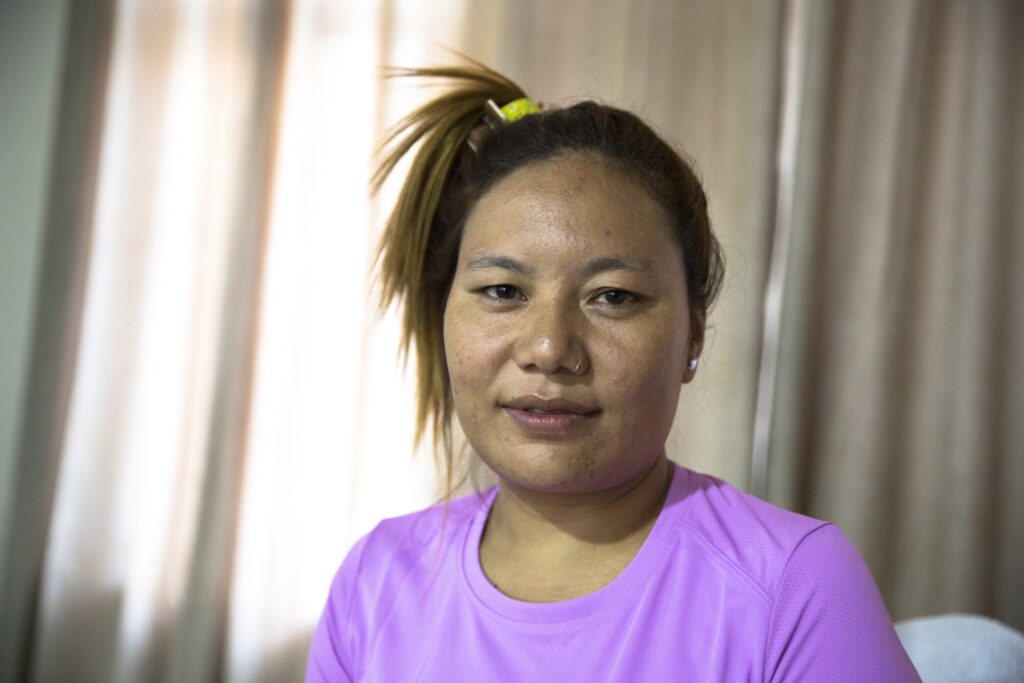 Portrait of Ganga. The young woman is wearing a side high ponytail. She has a nose piercing, is wearing a purple sports shirt and a yellow hair tie.