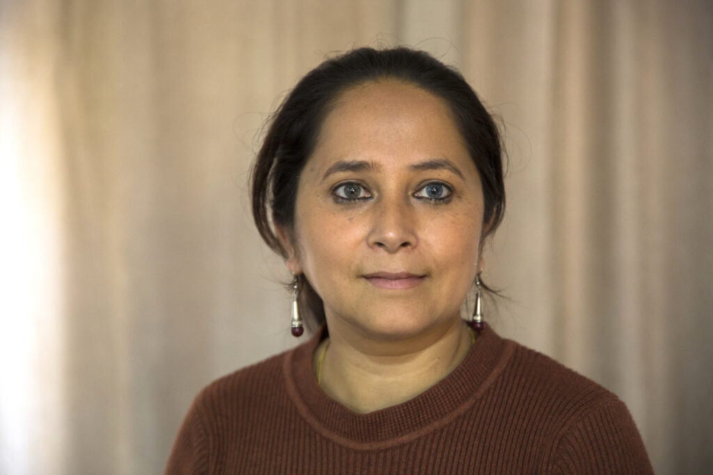 Portrait of Rashmi. Her hair is tied back, and her eyes are made up with kohl. She wears long silver earrings and a brown sweater.