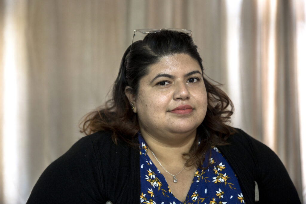 Portrait of Rupa. She is wearing glasses on the top of her head. She has a nose piercing and is wearing bright lipstick and a silver necklace. Rupa is a slightly overweight woman. She is wearing a black cardigan over a blue top with a bright floral pattern.