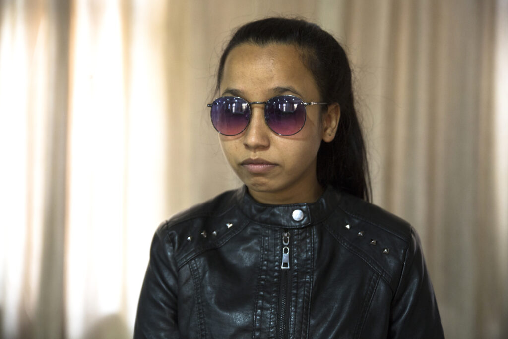 Portrait de Sunita N. Elle est jeune et a de longs cheveux noirs attachés en queue de cheval. Elle porte des lunettes de soleil rondes avec des verres de couleur violette et une monture argentée. Elle porte une veste en cuir noir.