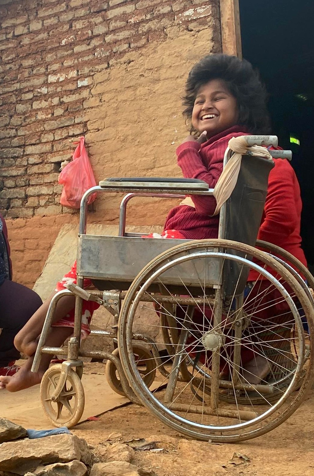 Una ragazza seduta su una sedia a rotelle. Sorride alla macchina fotografica. È seduta di fronte a una casa in mattoni.