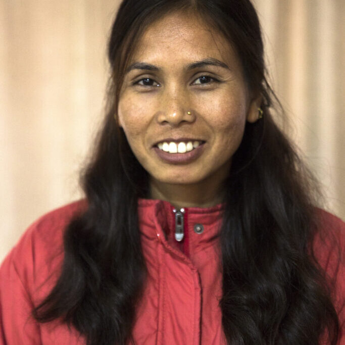 Portrait of Babita. Her hair drapes to the front of her shoulders. She is smiling, showing her white teeth. She has a small, silver nose piercing and is wearing a red jacket this is closed all the way to the top.