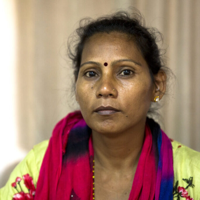 Portrait de Deu Kumari, qui regarde sérieusement dans l'appareil photo. Elle porte les cheveux attachés, de petites boucles d'oreilles, un piercing au nez et un bindi. Deu Kumari porte un foulard rose, bleue et noire et un débardeur jaune.