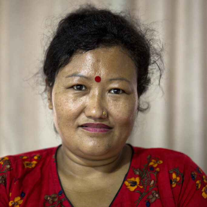 Portrait of Kamala. She has tied back her dark curls. She wears a bindi on her forehead, a nose piercing and lipstick. Her red dress has a floral pattern.